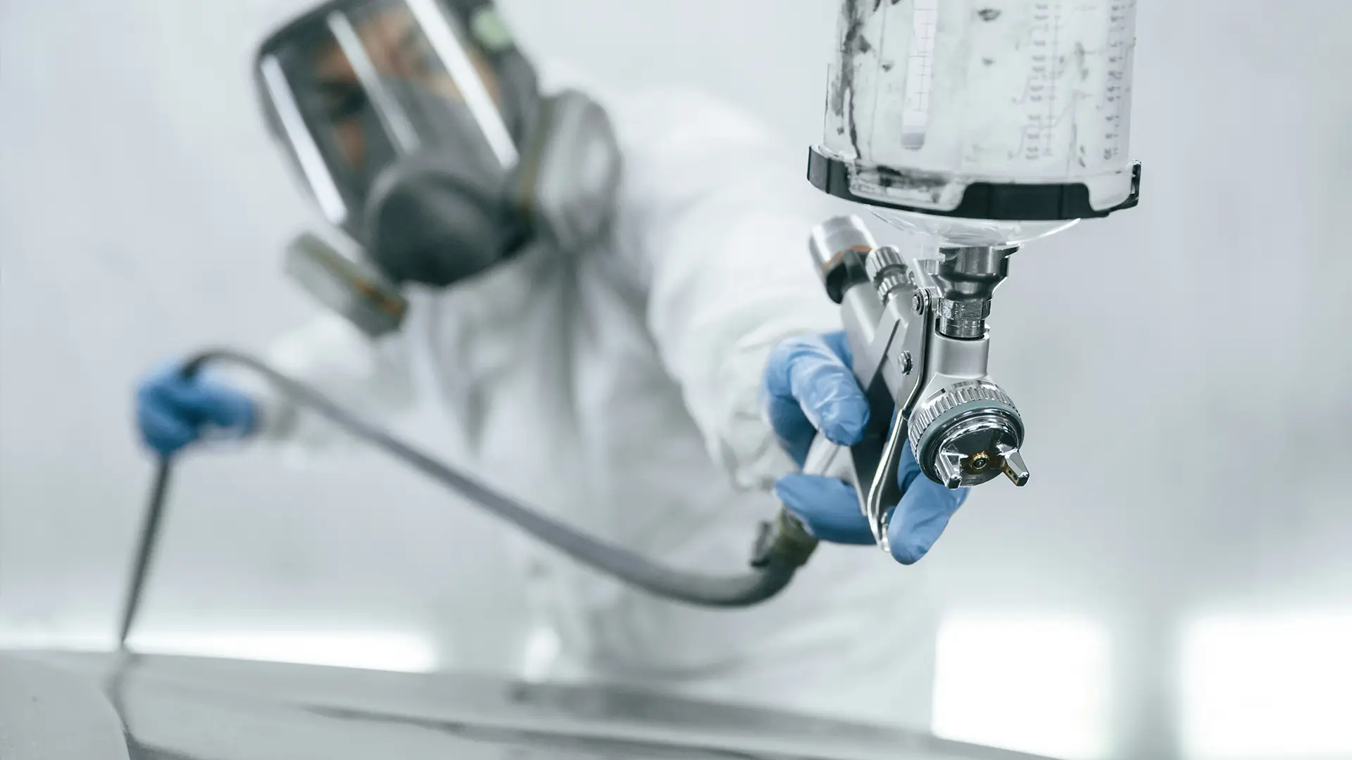 Close-up of a paint spray gun in focus with a technician in full PPE blurred in the background